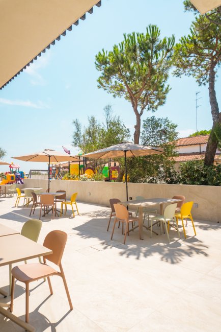 Area esterna con tavolini e sedie sotto ombrelloni e giochi per bambini sullo sfondo.