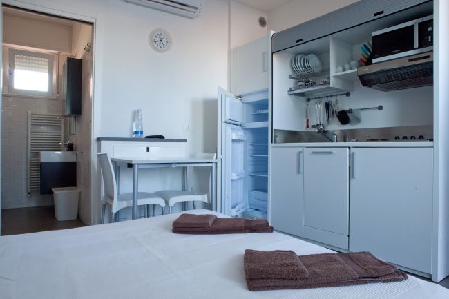 Interno angolo cottura con letto, tavolo bianco e accesso a un bagno.