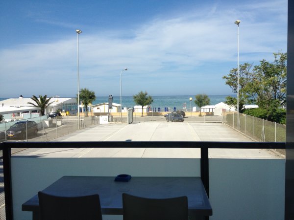Vista sul mare da un balcone, con auto parcheggiate e cielo blu.