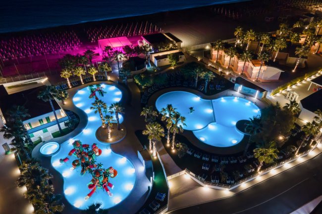 Piscine illuminate di notte con palme e giochi d'acqua, spiaggia sullo sfondo.