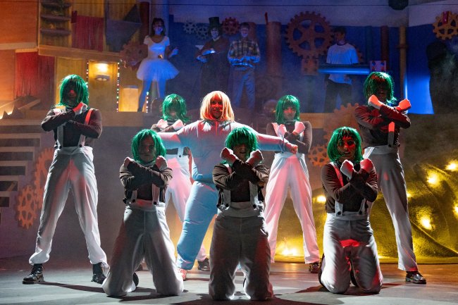 Persone con parrucche verdi e tute bianche eseguono una coreografia su un palco teatrale.