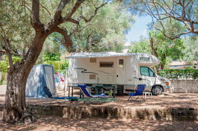 Camper parcheggiato tra alberi con sedie e tavolo fuori.