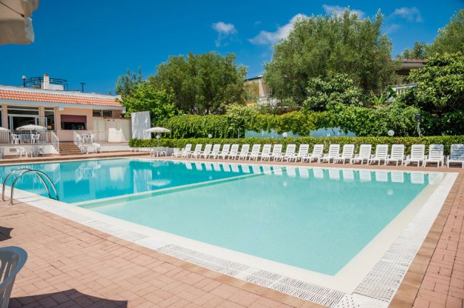 Piscina con lettini bianchi e ombrelloni, immersa nel verde e con cielo blu.
