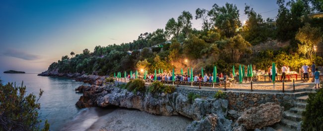 Tavolo lungo all'aperto su una terrazza rocciosa, illuminato al tramonto vicino al mare.