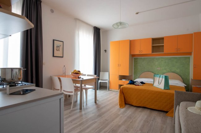 Un monolocale con angolo cottura, letto matrimoniale arancione, tavolo da pranzo e armadi arancioni.