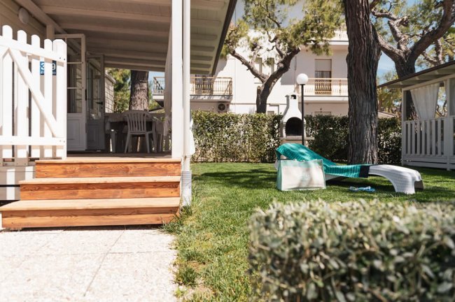 Porticato in legno con tavolo e sedie bianche. Giardino verde vicino a una siepe e un edificio.