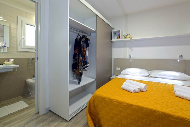 Camera con letto matrimoniale, coperta arancione, asciugamani, armadio aperto e bagno a vista.