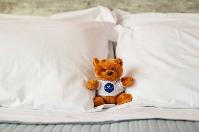 Orsetto di peluche su cuscini bianchi, indossa una maglietta con logo blu.