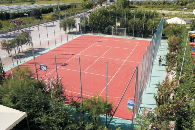 Campo sportivo recintato con superficie rossa, circondato da alberi e aree verdi.