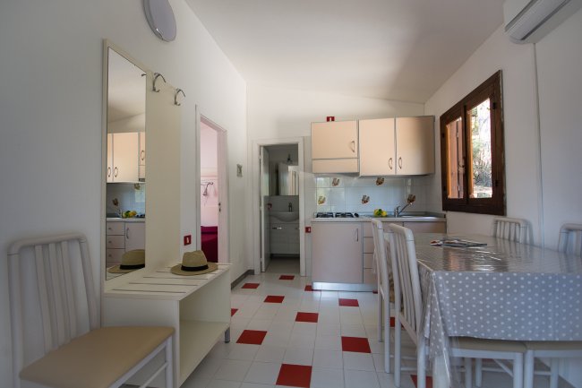 Cucina con tavolo da pranzo, sedie bianche e pensili beige. Pavimento bianco con piastrelle rosse.