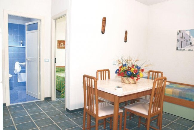 Sala da pranzo con tavolo, sedie e fiori. Porta aperta su bagno blu.
