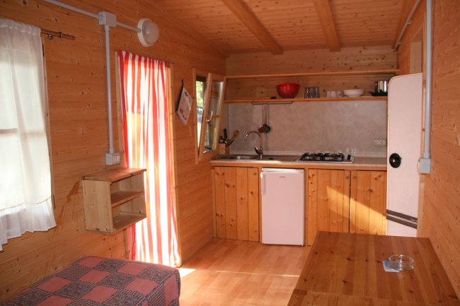 Interno di una casetta in legno con cucina compatta, frigorifero e tavolo.