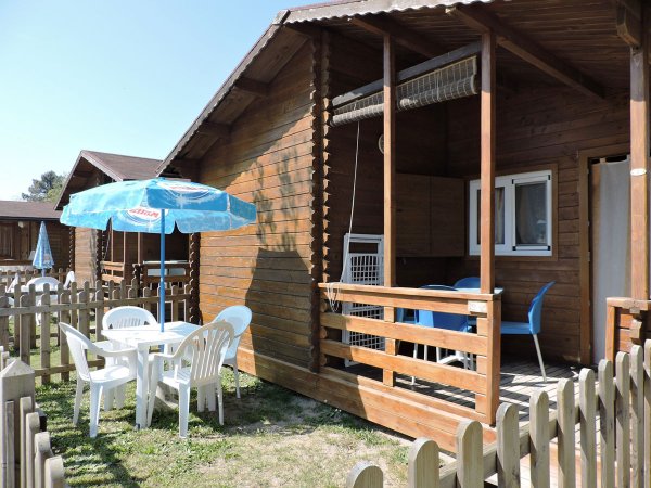 Chalet in legno con terrazza coperta, tavolo blu, sedie e ombrellone azzurro in giardino.