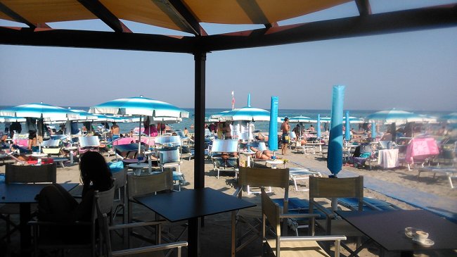 Vista dalla terrazza di un bar sulla spiaggia con ombrelloni e lettini blu.