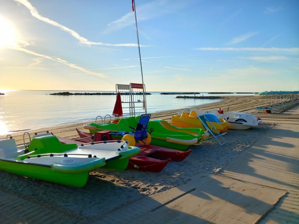 Pedalò colorati sulla sabbia al tramonto con mare calmo e cielo sereno.