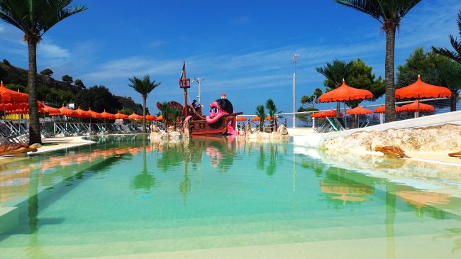 Piscina bassa con scenografie di pirati e ombrelloni arancioni sullo sfondo.