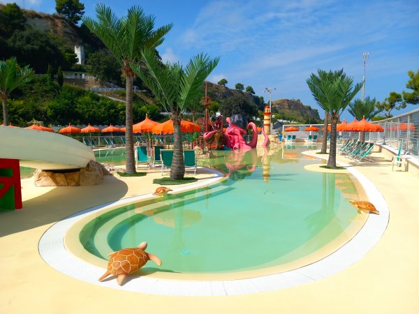 Piscina con scivoli, palme e decorazioni a tema tartaruga, ombrelloni arancioni sullo sfondo.