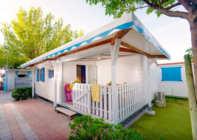 Una casetta bianca con veranda e tende blu, circondata da alberi e prato.