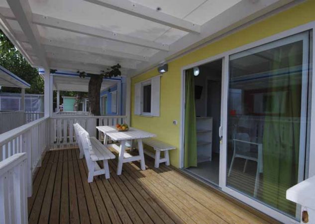 Veranda in legno con tavolo e panche bianche, vicino a una porta scorrevole in vetro.