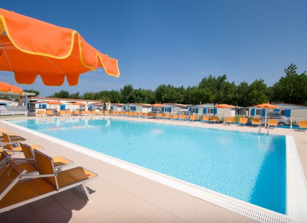 Piscina vuota con lettini e ombrelloni arancioni, cielo sereno e alberi sullo sfondo.