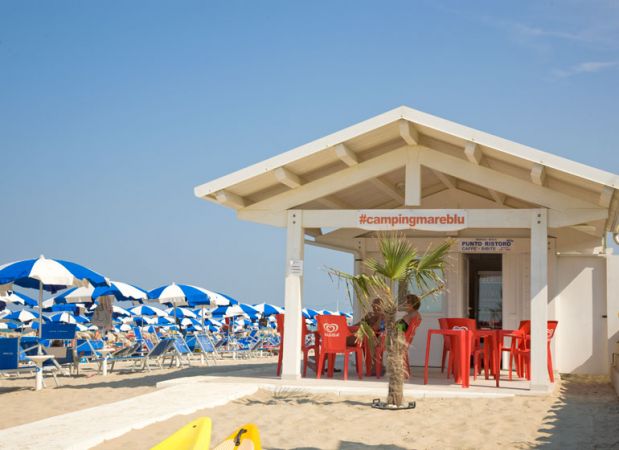 Chiosco bianco sulla spiaggia con sedie rosse. Ombrelloni blu e bianchi sullo sfondo.