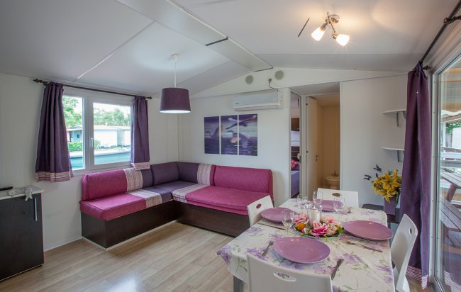Interno di una casa con divano angolare viola, tavolo apparecchiato e cucina a vista.