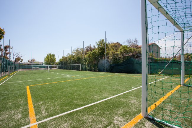 Un campo di calcio con erba sintetica e linee gialle, recintato, con alberi intorno e cielo sereno.