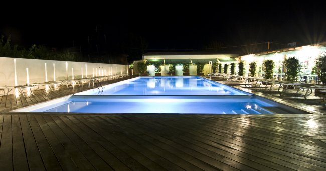 Una piscina illuminata di notte con lettini su una terrazza in legno.