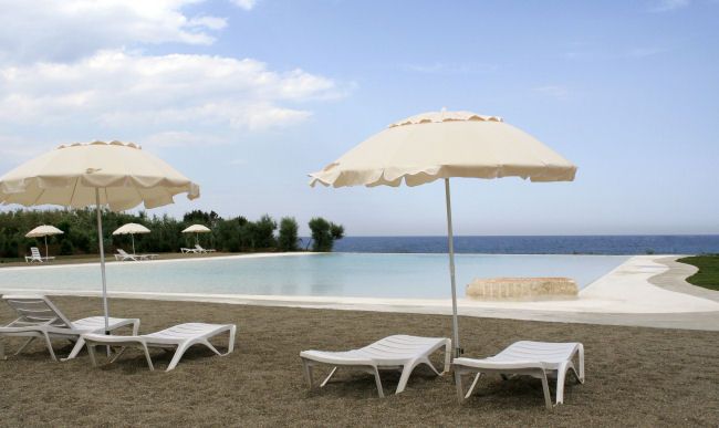 Due lettini bianchi sotto ombrelloni beige vicino a una piscina con vista mare.