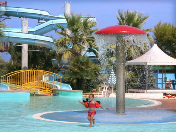 Bambina con braccioli in piscina, sotto un fungo a pioggia, vicino a scivoli d'acqua e palme.