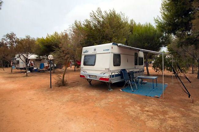 Camper parcheggiato su terreno arido, circondato da alberi e sedie pieghevoli sotto una tenda.