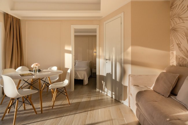 Soggiorno elegante con tavolo rotondo, sedie bianche e divano beige vicino alla porta della camera.