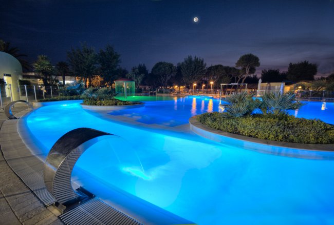 Piscina luminosa di notte con getti d'acqua e piante, sotto un cielo stellato.