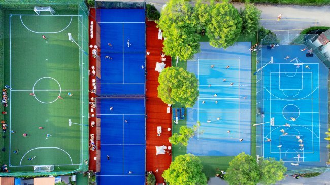 Vista dall'alto di campi sportivi colorati: calcio, paddle e basket, circondati da alberi.