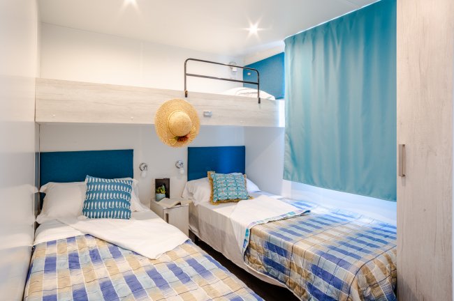 Camera con letti singoli, copriletti a righe, tenda blu e cappello appeso.