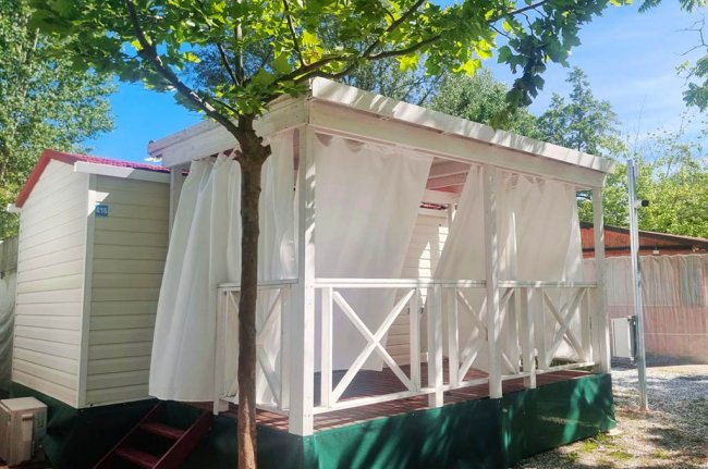 Un bungalow bianco con tende su una veranda, circondato da alberi.
