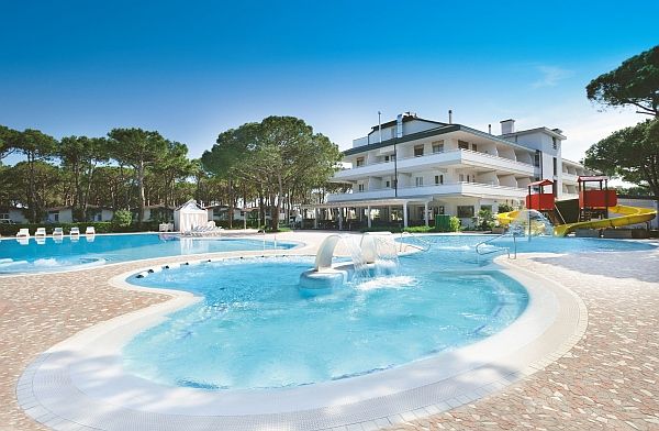 Una piscina con fontane, scivoli gialli e un edificio bianco sullo sfondo, circondato da alberi.